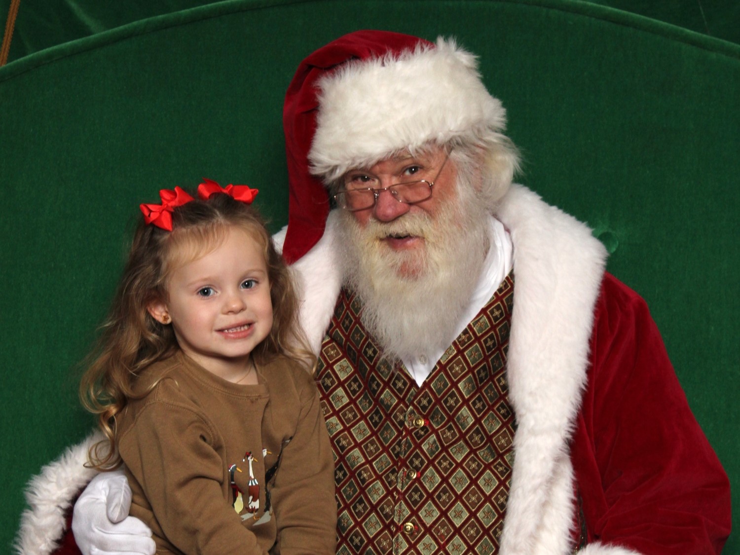 A young child in a festive sweater sits on Santa's lap, both smiling against a green backdrop, capturing a cheerful holiday moment.